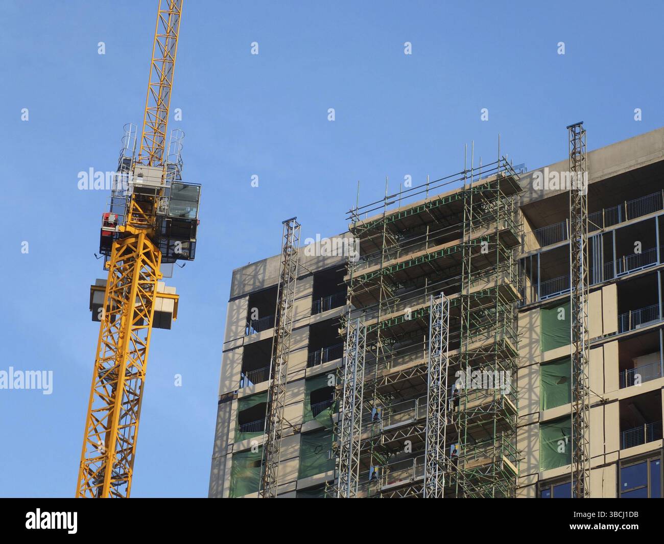 Ein gelber Baukran neben einem großen Betongebäude im Bau mit Gerüsten und Hubtürmen Stockfoto