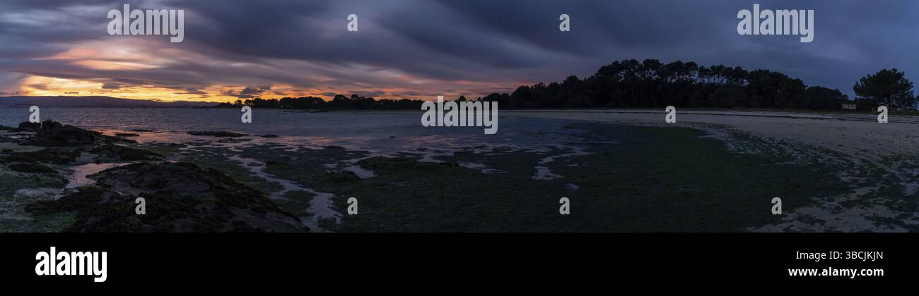 Ein Panoramablick auf einen Strand bei Ebbe unter Ein bedecktes Nachthimmel mit Tagesanbruch und buntem Sonnenaufgang Stockfoto
