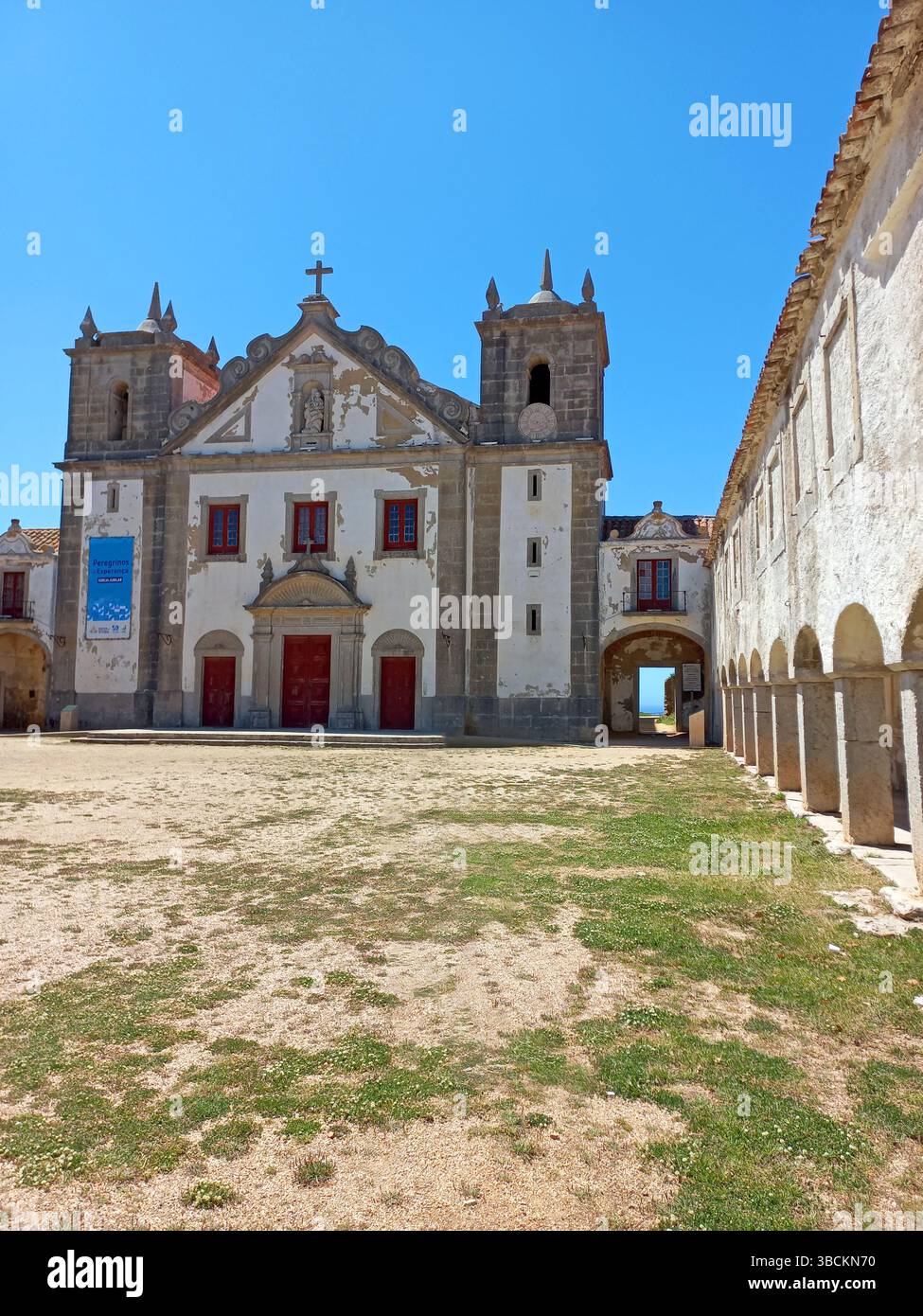 Altes religiöses Heiligtum am Kap Espichel in Sesimbra Portugal mit langen symmetrischen Arkaden und einer historischen Kirchenfassade unter einem leuchtenden Blau Stockfoto