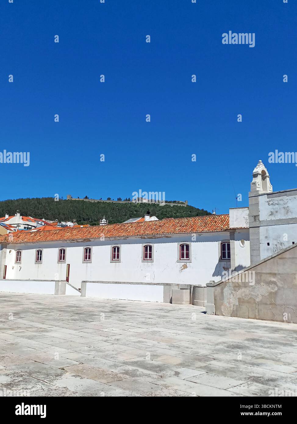Traditionelle weiße Gebäude mit roten Dächern im Inneren der Festung Sesimbra mit Blick auf eine ferne Burg und bewaldete Hügel unter klarem Himmel Stockfoto