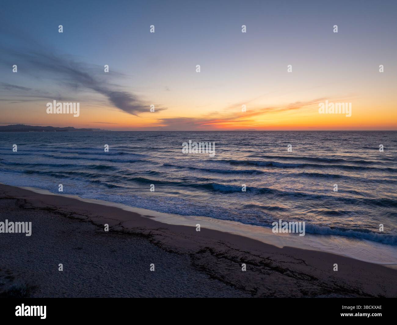 Ruhiger Sonnenuntergang am Strand mit sanften Wellen, die unter einem leuchtenden orange-blauen Himmel an Land ziehen. Stockfoto