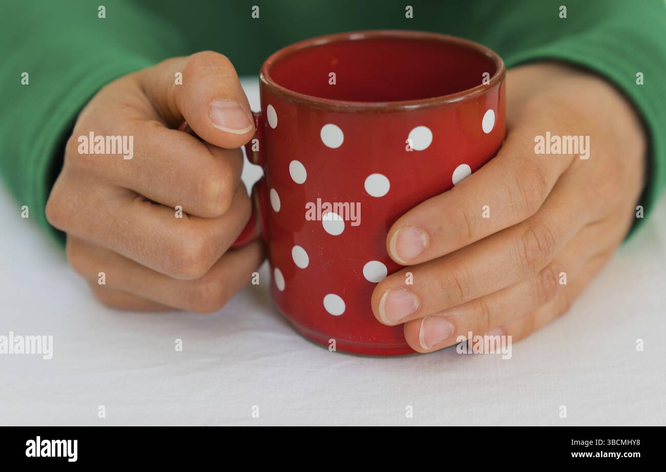 Zarte weibliche Hände einer Frau in einem hellen grünen Pullover holding Eine leere Kaffeetasse Rot mit weißen Tupfen auf einer weißen Tischdecke Stockfoto