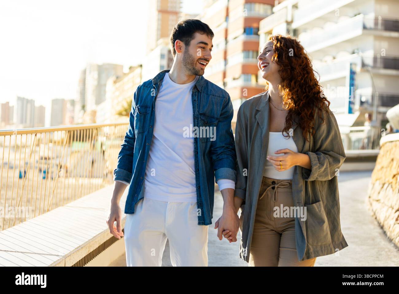 Lächelndes junges Paar spaziert Hand in Hand auf einer sonnigen urbanen Promenade und teilt Lachen und Gespräche mit modernen Gebäuden und Strand im Hinterland Stockfoto
