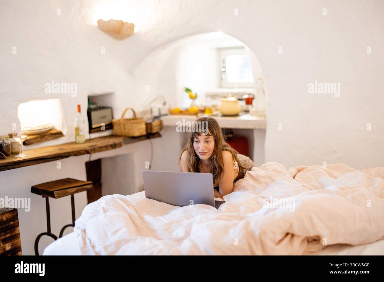 Eine junge Frau liegt auf einem gemütlichen Bett mit einem Laptop in einem weiß getünchten trullo Haus. Das rustikale Interieur, die Holzregale und das warme Licht schaffen eine ruhige, abgelegene Arbeitsszene in Süditalien Stockfoto