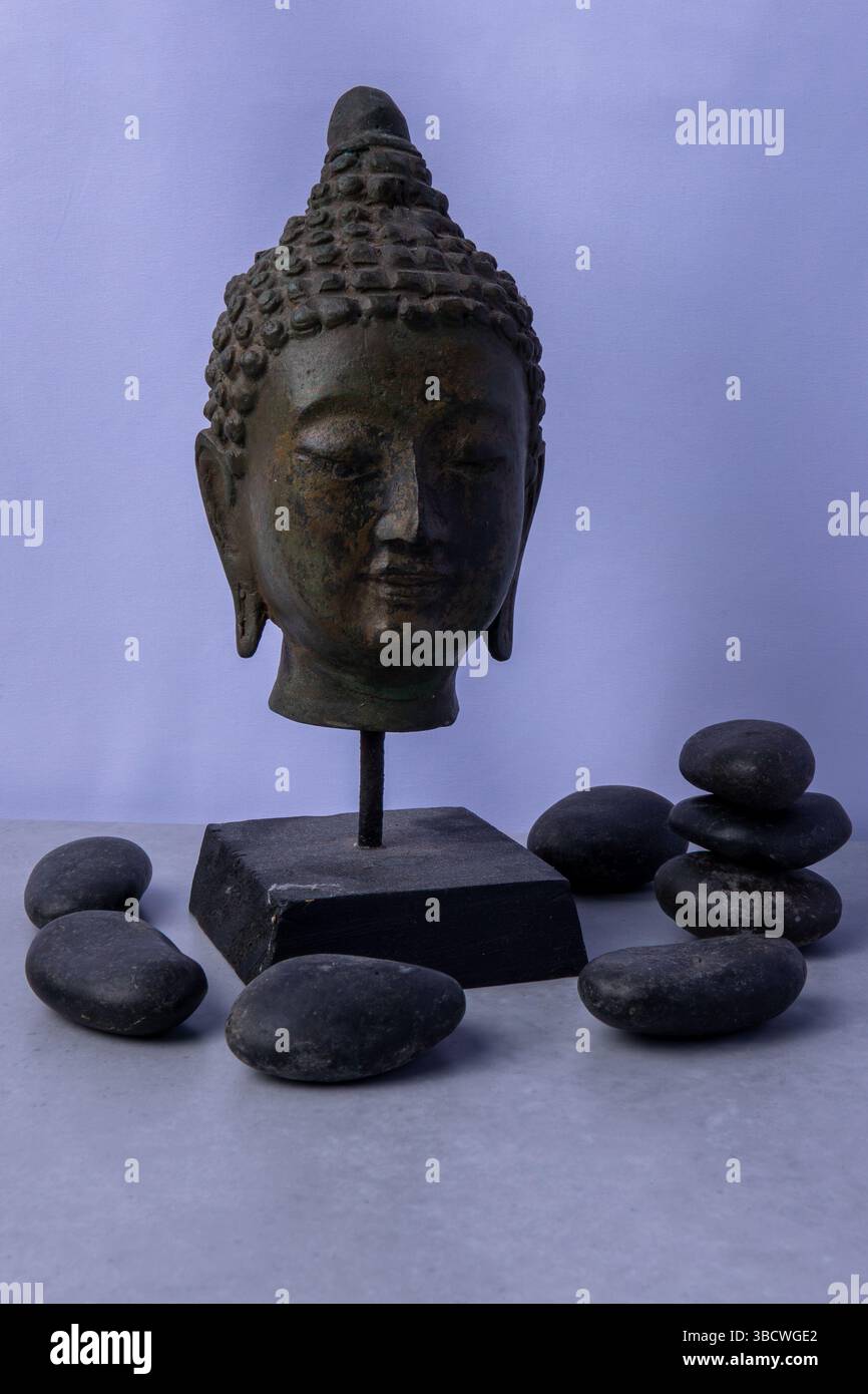 Die Skulptur des Buddha-Kopfes steht auf einem Sockel, umgeben von glatten schwarzen Steinen und bietet eine beruhigende und friedliche Atmosphäre. Stockfoto