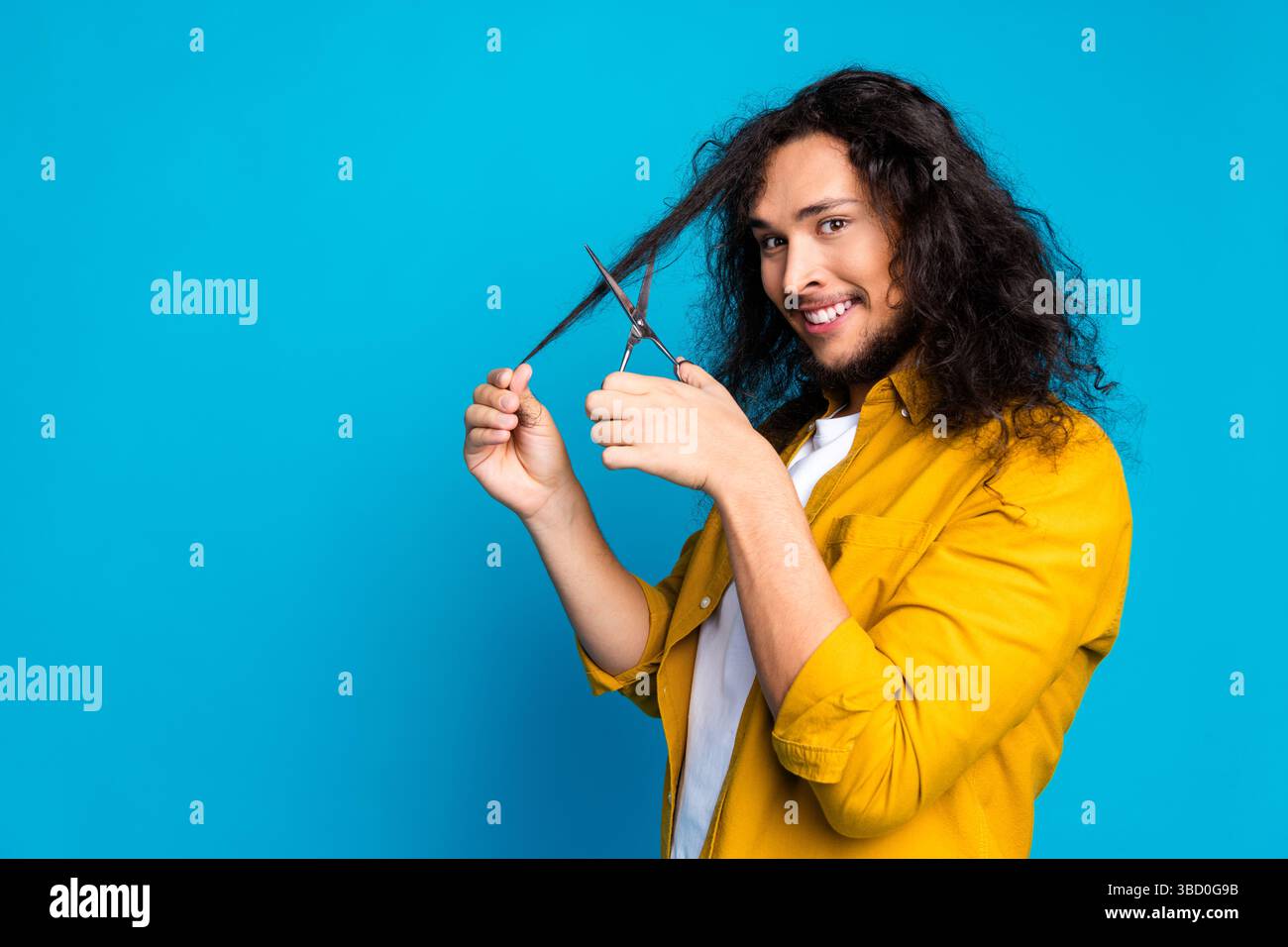Selbstbewusster junger Mann trimmt sein langes lockiges Haar mit einer Schere vor einem hellblauen Hintergrund und lächelt fröhlich Stockfoto