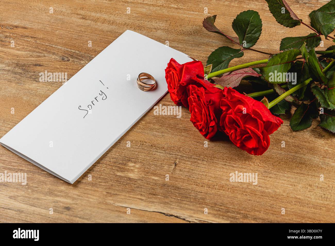 Eine handschriftliche Notiz mit der Aufschrift "Sorry" liegt neben einem Ehering und einem Strauß roter Rosen auf einem rustikalen Holztisch, was eine herzliche Entschuldigung symbolisiert Stockfoto