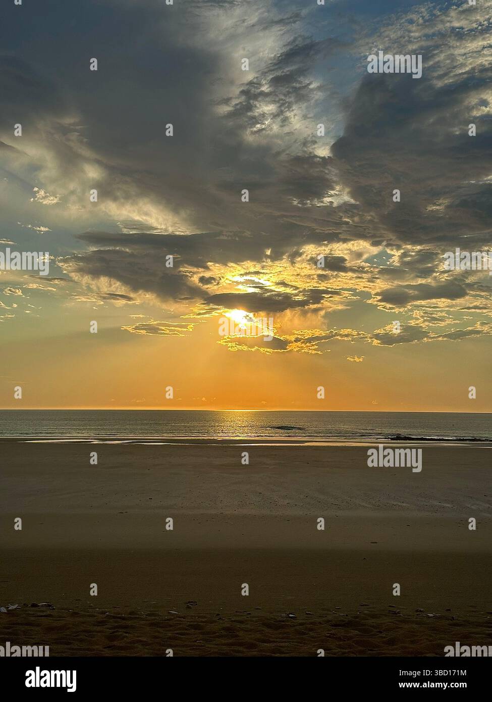 Ruhiger Sonnenaufgang über einem ruhigen, leeren Strand mit goldenem Sonnenlicht, das auf ruhigem Meereswasser reflektiert, und dramatischen Wolkenformationen am Himmel - Smartphone-aufgenommenes Stockfoto