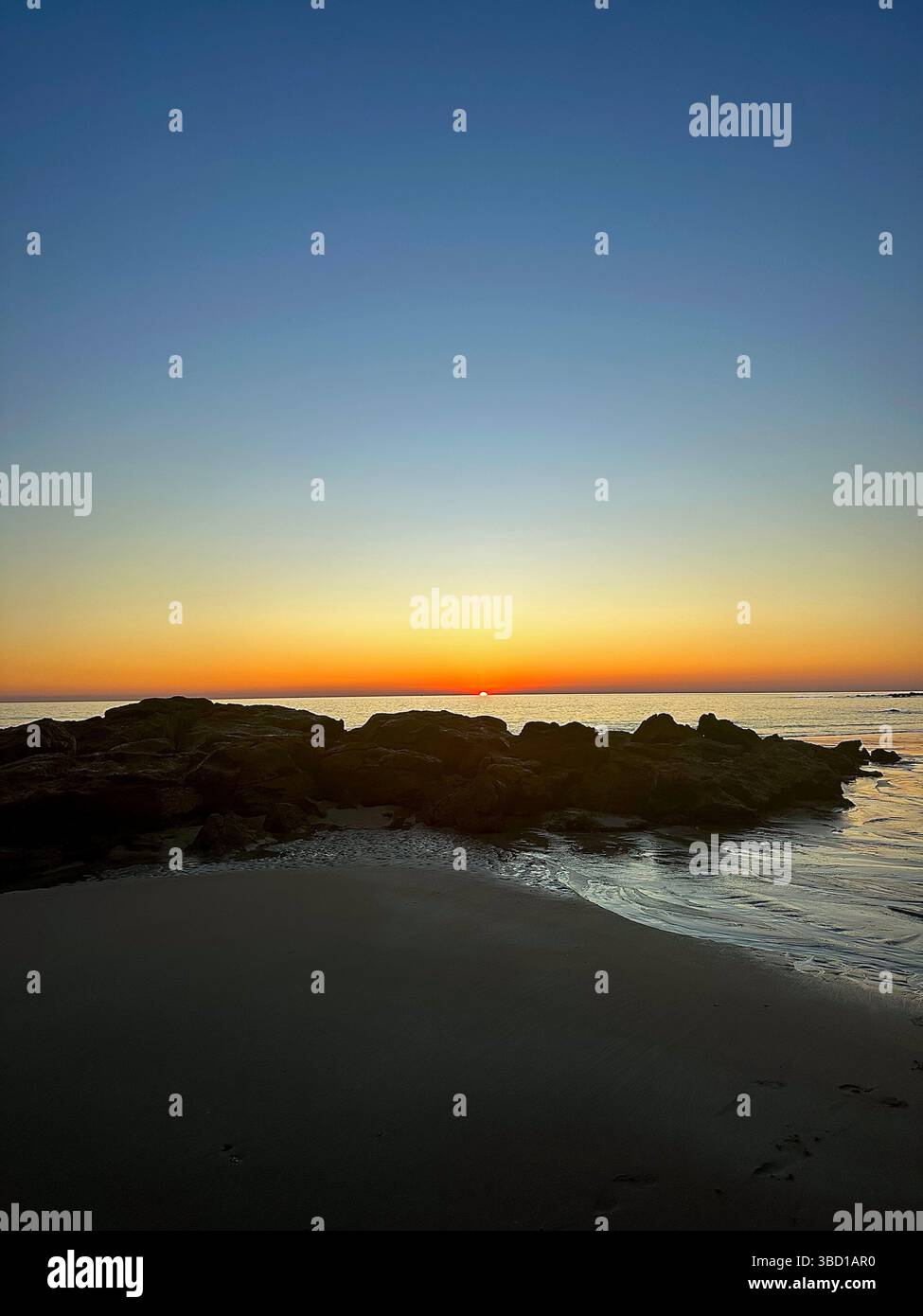 Ruhiger Sonnenaufgang am Strand mit Silhouette-Felsen und sanften Reflexen auf nassem Sand – perfekt für Wellness, Reisen und inspirierende Themen. - Smartphone-aufgenommenes Stockfoto