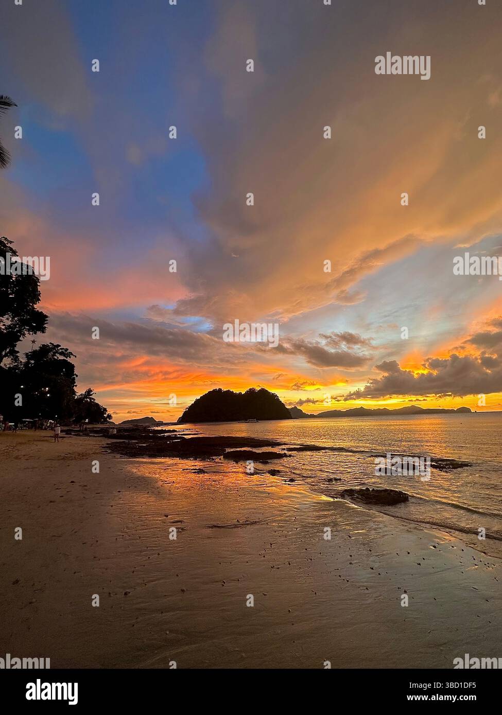 Pulsierender tropischer Sonnenuntergang über einem ruhigen Strand mit felsiger Küste und Inselsilhouette, die goldene Reflexe über das ruhige Meereswasser wirft - Smartphone-aufgenommenes Stockfoto
