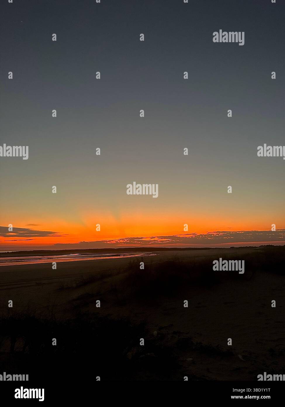 Dramatisches Licht vor der Dämmerung über dem Horizont des Ozeans, das bei Sonnenaufgang tiefe Orange- und Goldtöne über den ruhigen Strand wirft. - Smartphone-aufgenommenes Stockfoto