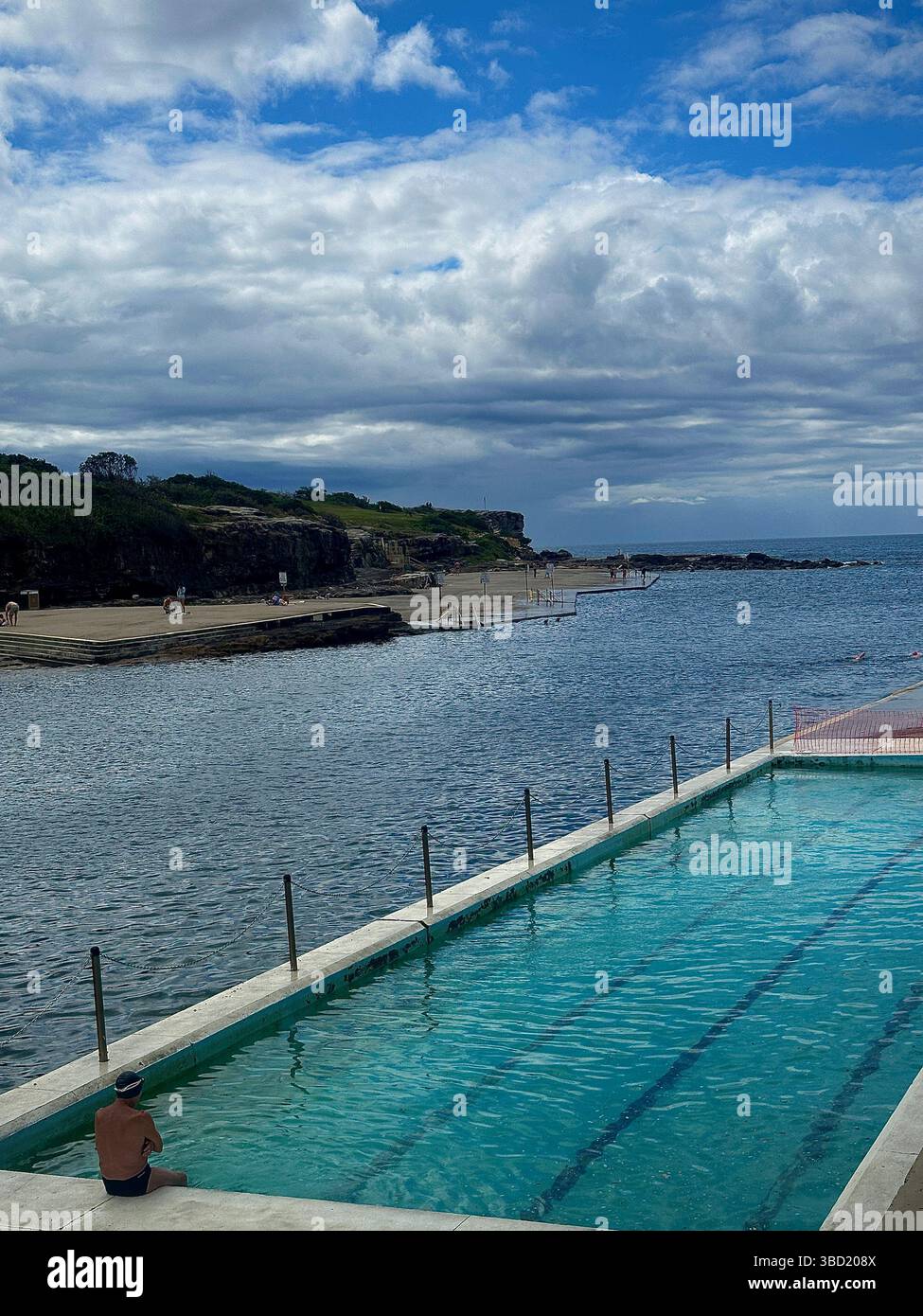 Ruhiger Pool mit Blick auf die felsige Küste und ruhiges, blaues Wasser unter teilweise bewölktem Himmel, bietet einen friedlichen Badeort. - Smartphone-aufgenommenes Stockfoto