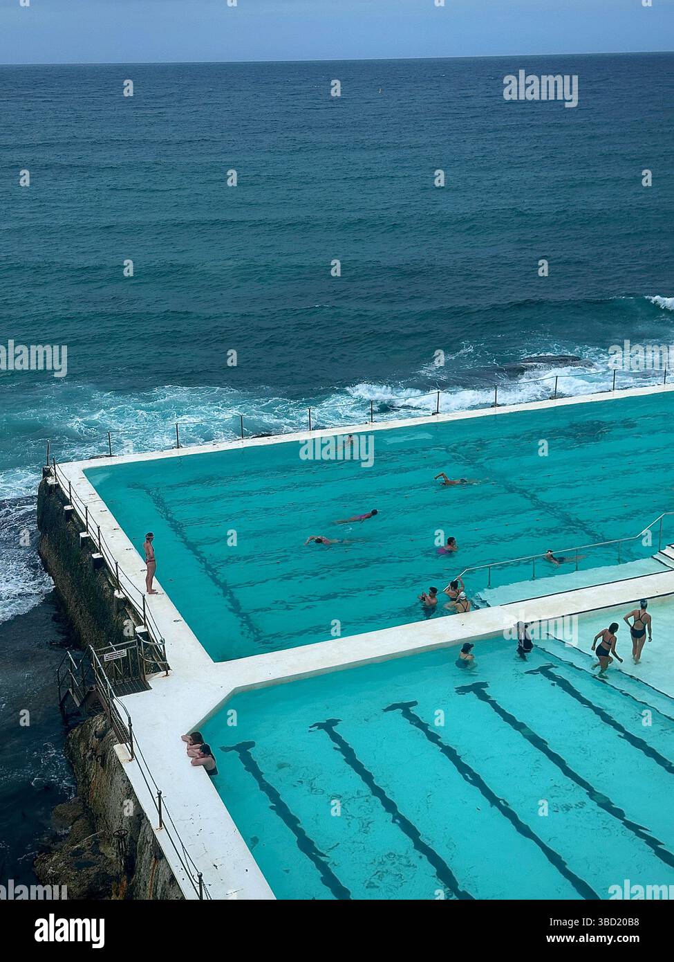 Schwimmer genießen den berühmten Bondi Icebergs Ozean Pool, der dramatisch am Rand der Küste thront, mit Wellen, die unten krachen. - Smartphone-aufgenommenes Stockfoto