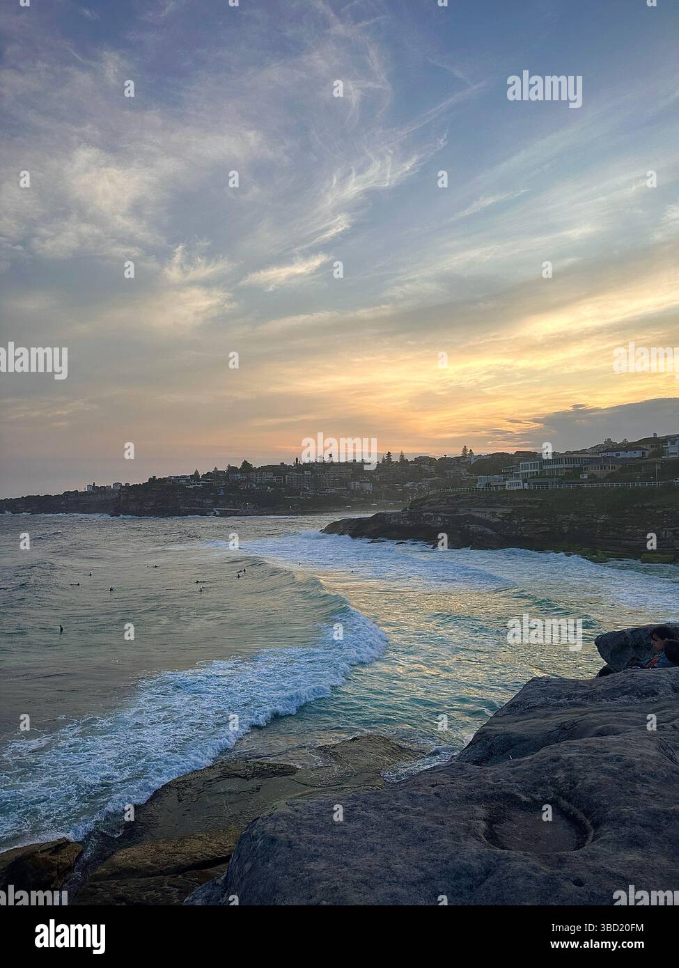 Goldener Sonnenuntergang über einer zerklüfteten australischen Küste mit sanften Wellen, die zur Küste Rollen und Surfer das Abendlicht genießen. - Smartphone-aufgenommenes Stockfoto
