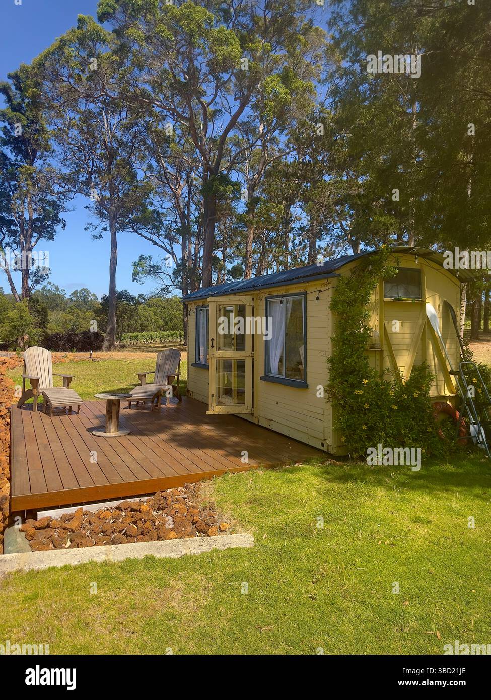 Charmantes, winziges Haus mit Holzterrasse in einer ruhigen Landschaft, umgeben von hohen Bäumen und üppig grünem Rasen, perfekt für einen entspannenden Urlaub auf dem Land - Smartphone-aufgenommenes Stockfoto