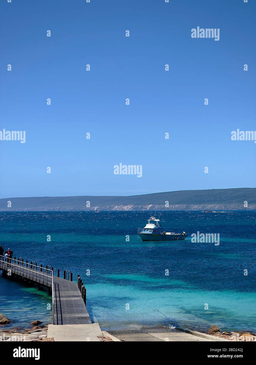 Idyllische Küstenszene mit einem hölzernen Pier, der zu einem Fischerboot führt, das in lebhaftem türkisfarbenem Wasser vor Anker liegt, eingerahmt von einem klaren blauen Himmel und einer fernen Küste - Smartphone-aufgenommenes Stockfoto