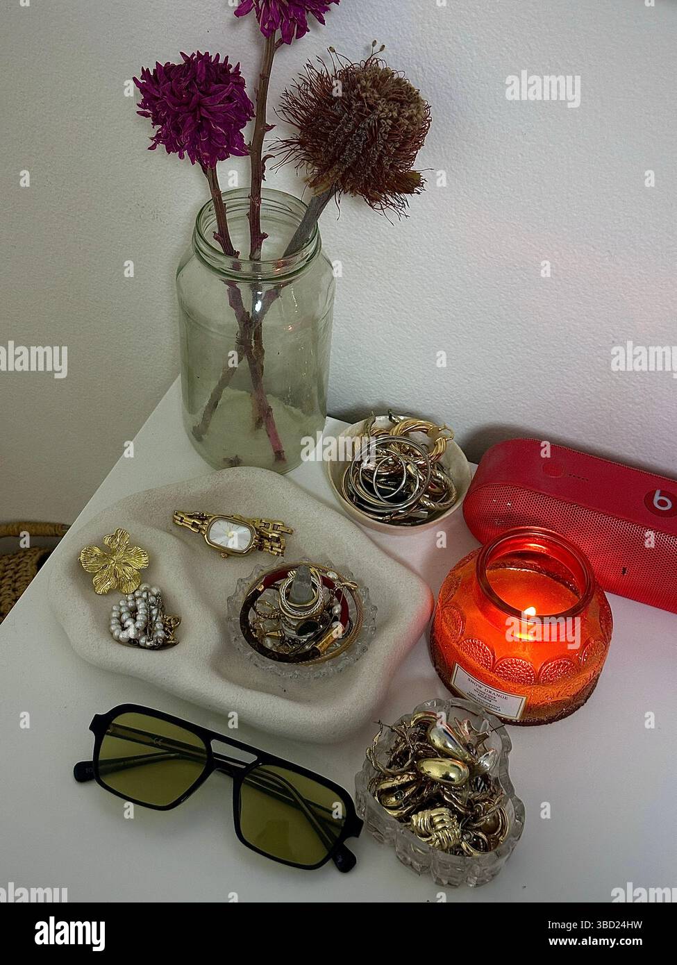 Gemütlicher Nachttisch mit Schmuckständer, dekorativen Blumen und einer leuchtenden Kerze für eine warme, einladende Atmosphäre. - Smartphone-aufgenommenes Stockfoto