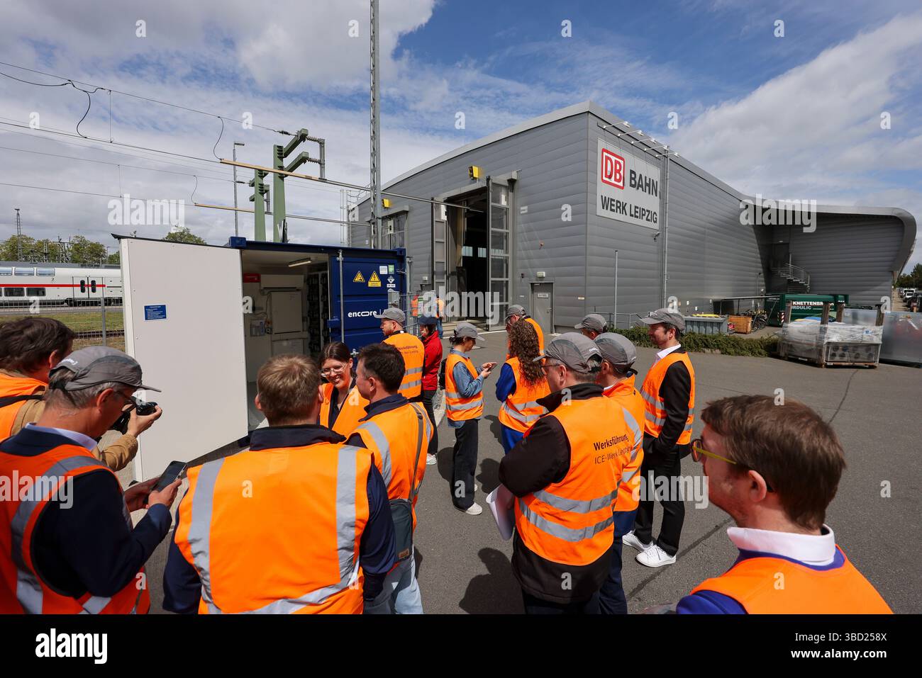 22. Mai 2025, Sachsen, Leipzig: Journalisten, Mitarbeiter und Gäste der Deutschen Bahn stehen vor dem neuen Stromspeicher des ICE-Werks Leipzig. Das sogenannte Second-Life-Batteriespeichersystem besteht aus alten Elektroautos, die über das firmeneigene Solarsystem aufgeladen werden. Die Solaranlage und der Batteriespeicher könnten rund ein Viertel des Strombedarfs des Leipziger EISWERKES decken. Durch das Pilotprojekt würden jährlich rund 85.000 Euro an Energiekosten eingespart. Der nächste Schritt besteht darin, das Instandhaltungswerk in Kassel mit einer Second-Life-Batterie auszustatten Stockfoto