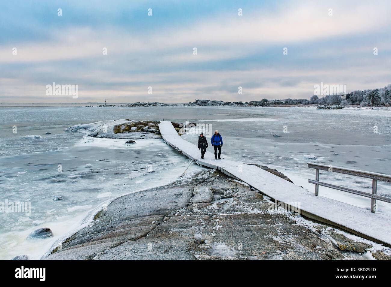 Zwei Personen schlendern entlang eines verschneiten, frostigen Weges, der zum Ufer der Insel Hönö an der schwedischen Küste führt. Die ruhige Landschaft wird hervorgehoben b Stockfoto