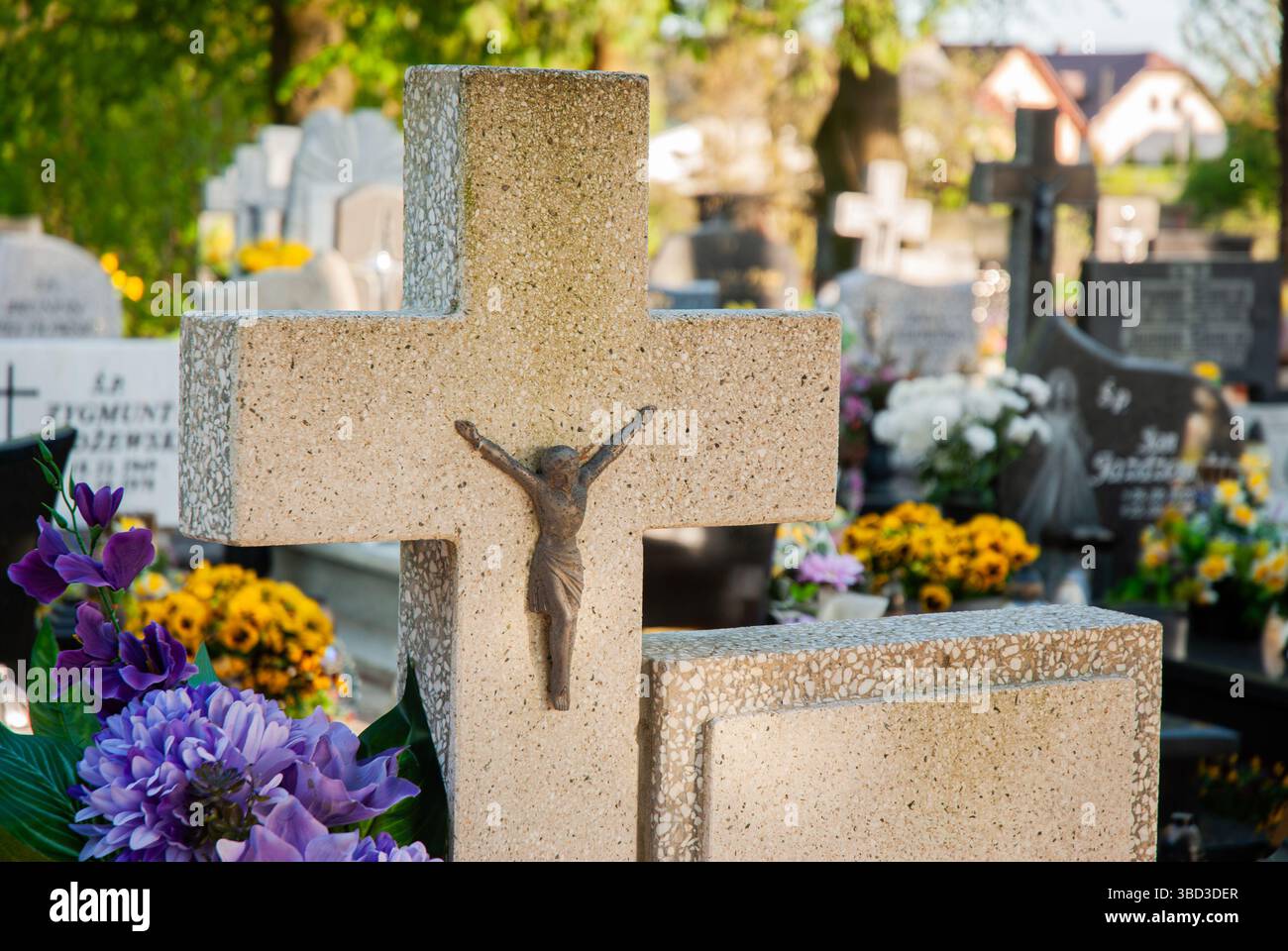 Das Steinkreuz mit einem Kruzifix steht inmitten lebendiger Blumen und Grabsteine auf einem friedlichen Friedhof unter einem sonnigen, klaren Himmel. Stockfoto