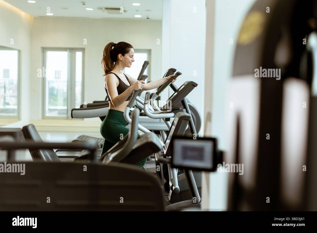 Eine Frau genießt ihre Fitness-Routine auf einem Crosstrainer, konzentriert und motiviert, umgeben von eleganten Fitnessgeräten bei hellem Tageslicht. Stockfoto