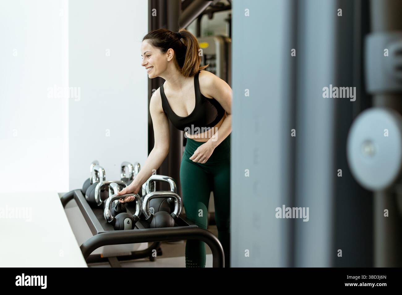 Eine junge Frau lächelt, als sie in einem stilvollen Fitnessstudio nach einer Kettlebell greift und ihre Leidenschaft für Fitness und Krafttraining zum Ausdruck bringt. Stockfoto