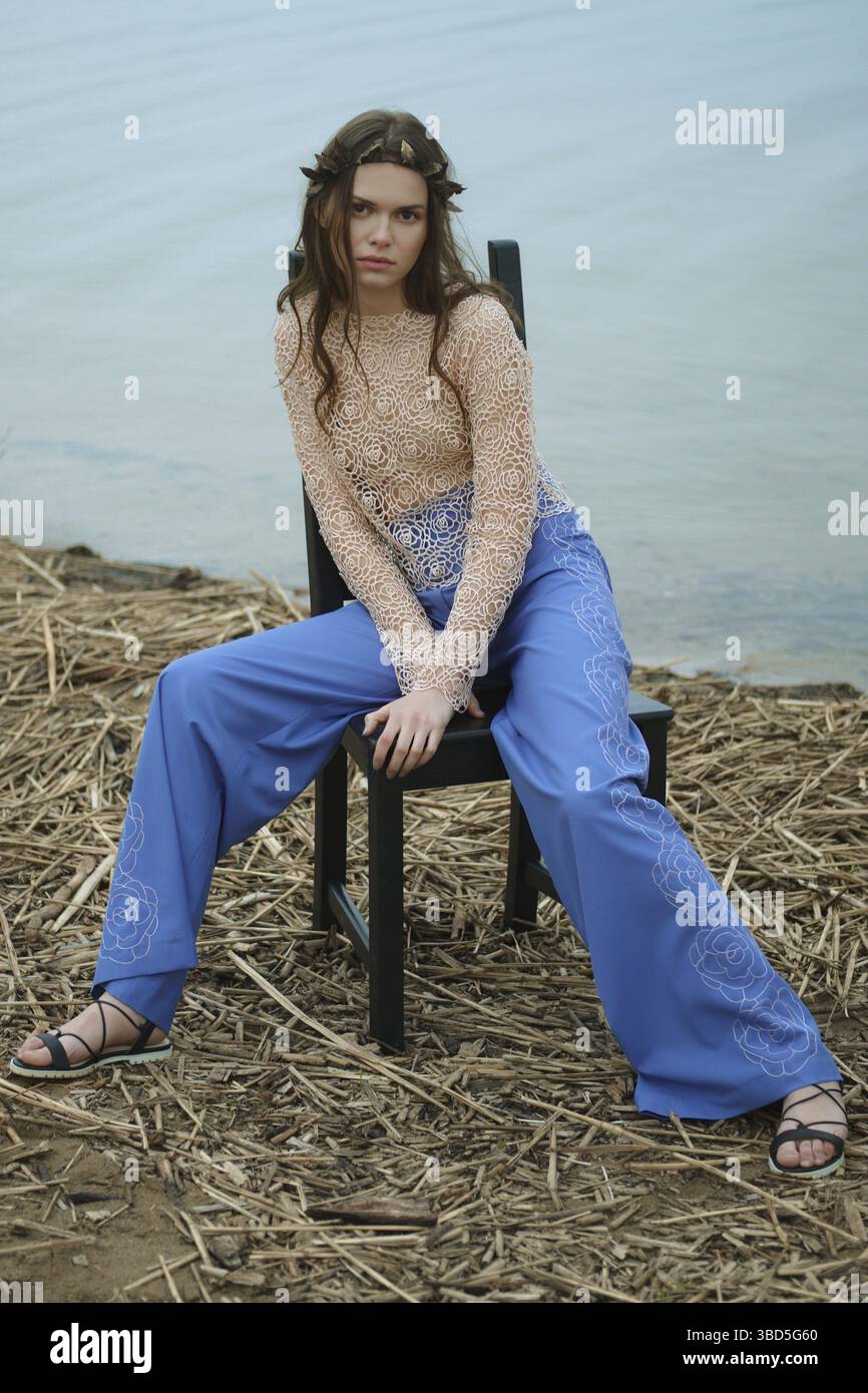 Porträt einer jungen Frau in weiten blauen Hosen und einer Spitzenbluse, die auf einem Stuhl am Ufer des Sees sitzt Stockfoto