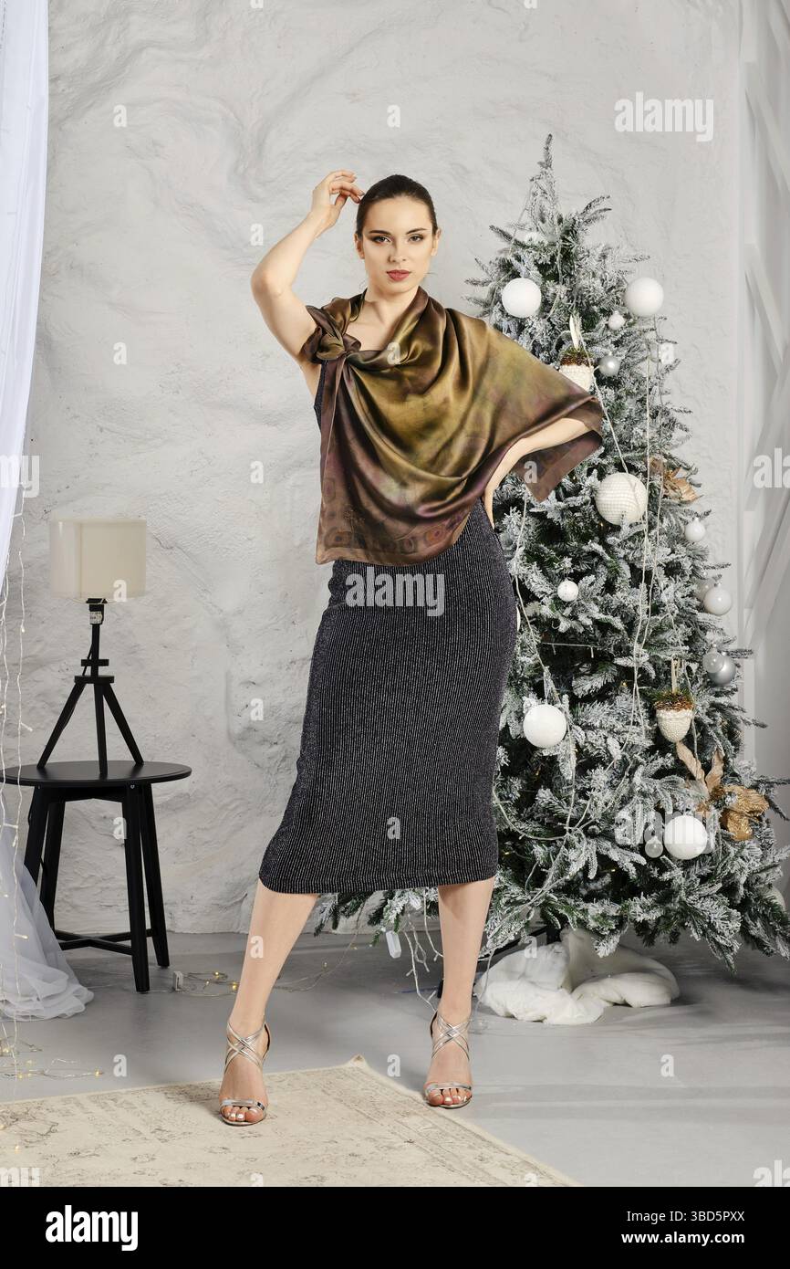 Eine junge Frau im engen schwarzen Kleid mit Glitzer und Seidenschal posiert neben dem Weihnachtsbaum Stockfoto