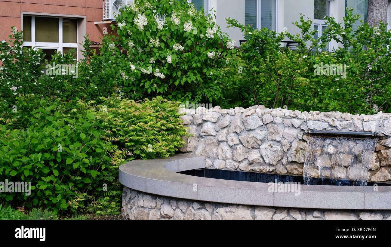 Dekorativer Teich mit Steinen in einem kleinen Garten. Blühender weißer Flieder. Kleiner Brunnen. Ein Innenhof in der Stadt. Stockfoto