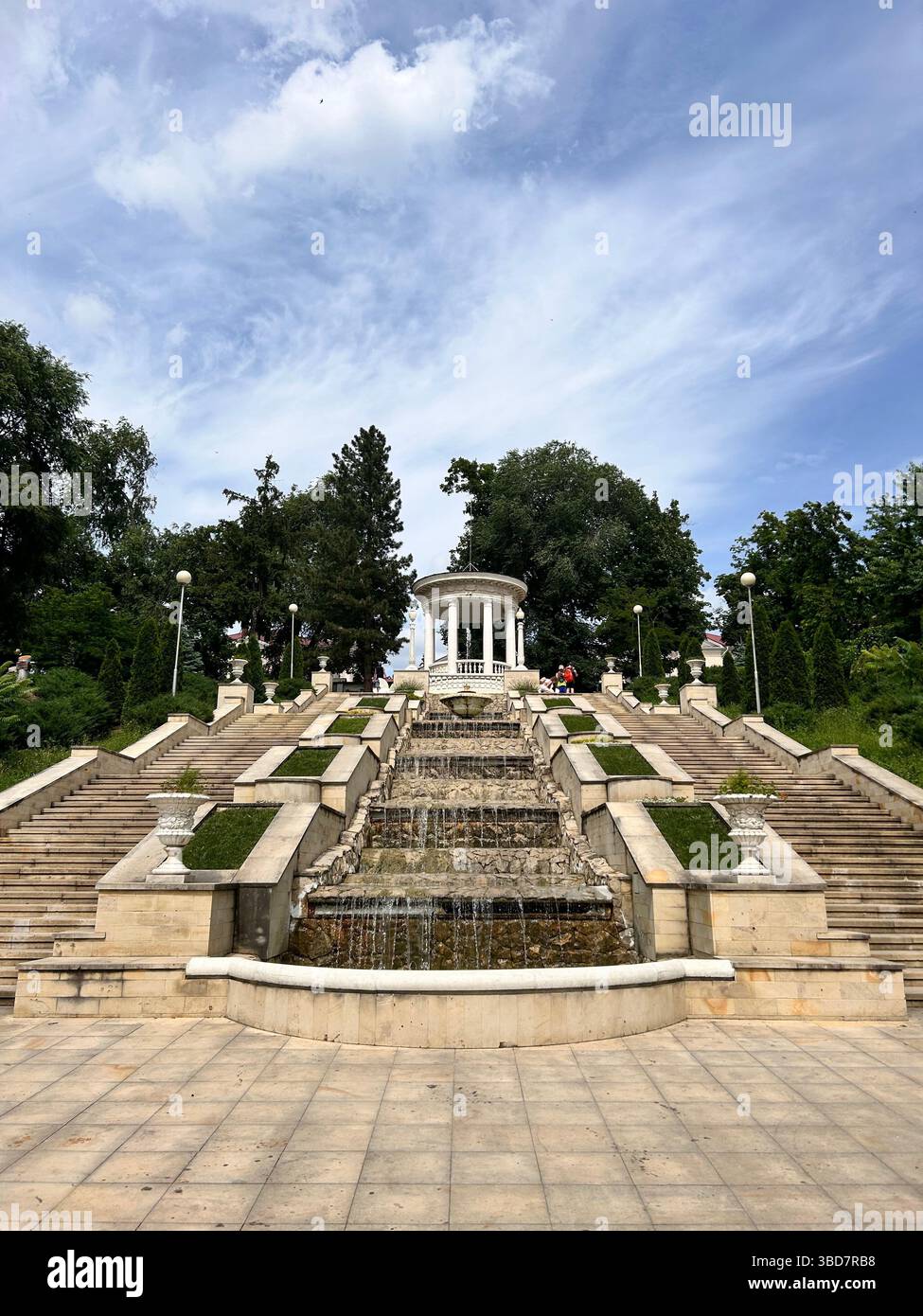 Der kaskadierende Steinbrunnen und die Treppe führen zu einer weißen Rotunde in einem landschaftlich gestalteten Park. Erschossen in Chisinau, Moldawien, Sommertag. - Smartphone-aufgenommenes Stockfoto
