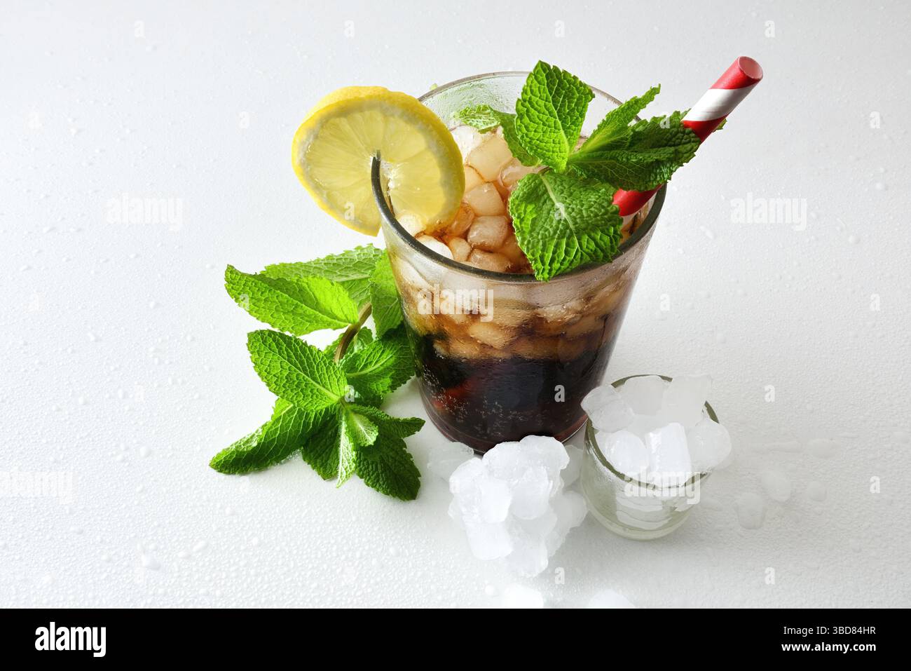Erfrischendes Cola-Getränk mit Eis in einer Glasschale, dekoriert mit einem Strohhalm und einer Scheibe Obst auf einem weißen Tisch mit Eis und Minzblättern. Erhöhte Aussicht. Stockfoto