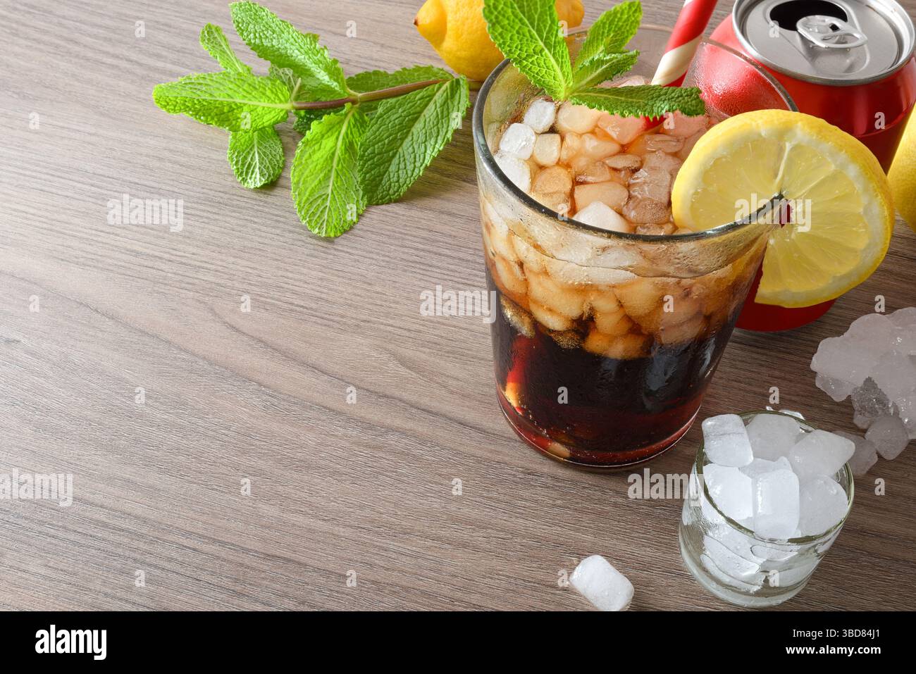 Cocktail auf Cola-Basis mit Minze und Zitrone in einem Glas auf einem Holztisch, dekoriert mit Eis, Minzblättern, Zitronen und einer Dose Cola. Erhöhte Aussicht. Stockfoto
