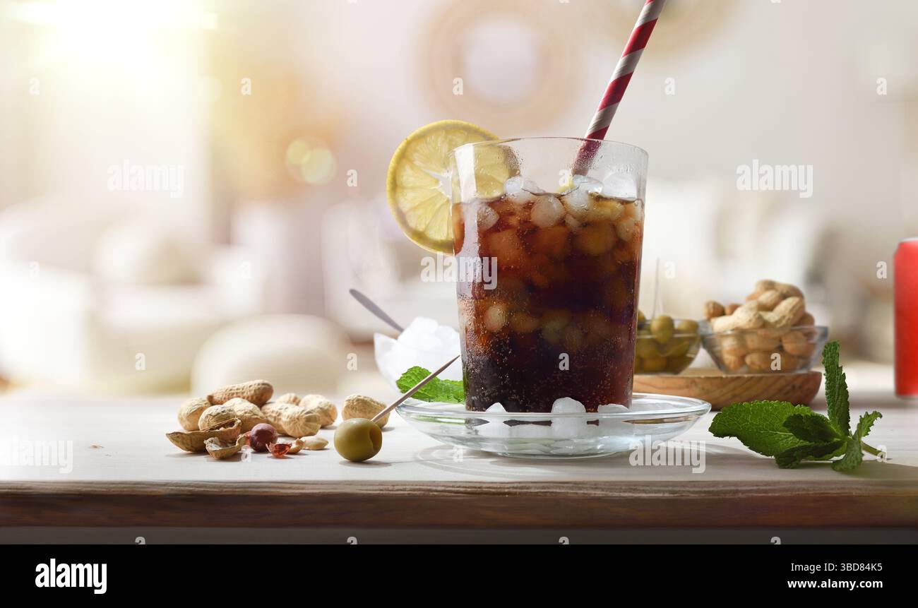 Erfrischendes Cola-Getränk auf einem Holztisch mit Snacks wie Erdnüssen und Oliven, dekoriert mit Minzblättern und Zitronenscheiben im Zimmer. Vorderansicht. Stockfoto