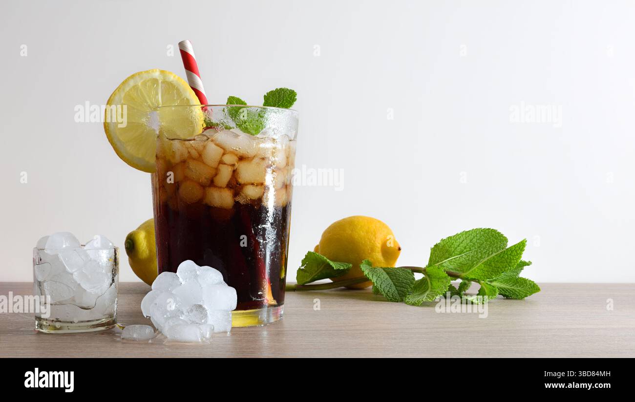 Erfrischendes Cola-Getränk mit Eis in einer Glasschale, dekoriert mit einem Strohhalm und einer Scheibe Obst auf einem Holztisch mit Obst, Eis und Minzblättern und einem Weiss Stockfoto