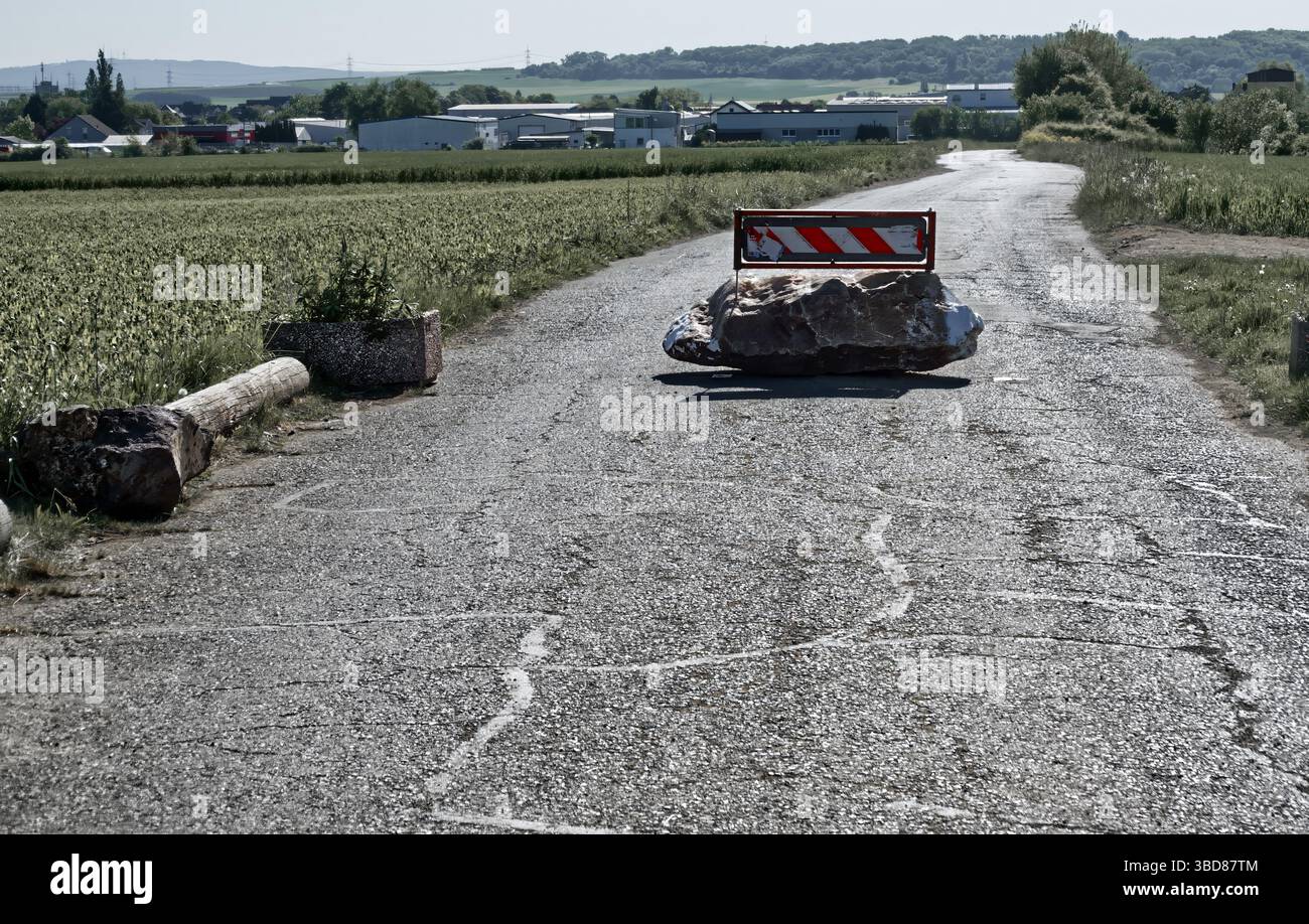 Die ländliche Straße ist durch einen großen Felsen und eine rot gestreifte Barriere blockiert, die vor einem Hintergrund von Feldern und fernen Hügeln liegt. Die Szene zeigt eine ungewöhnliche m Stockfoto