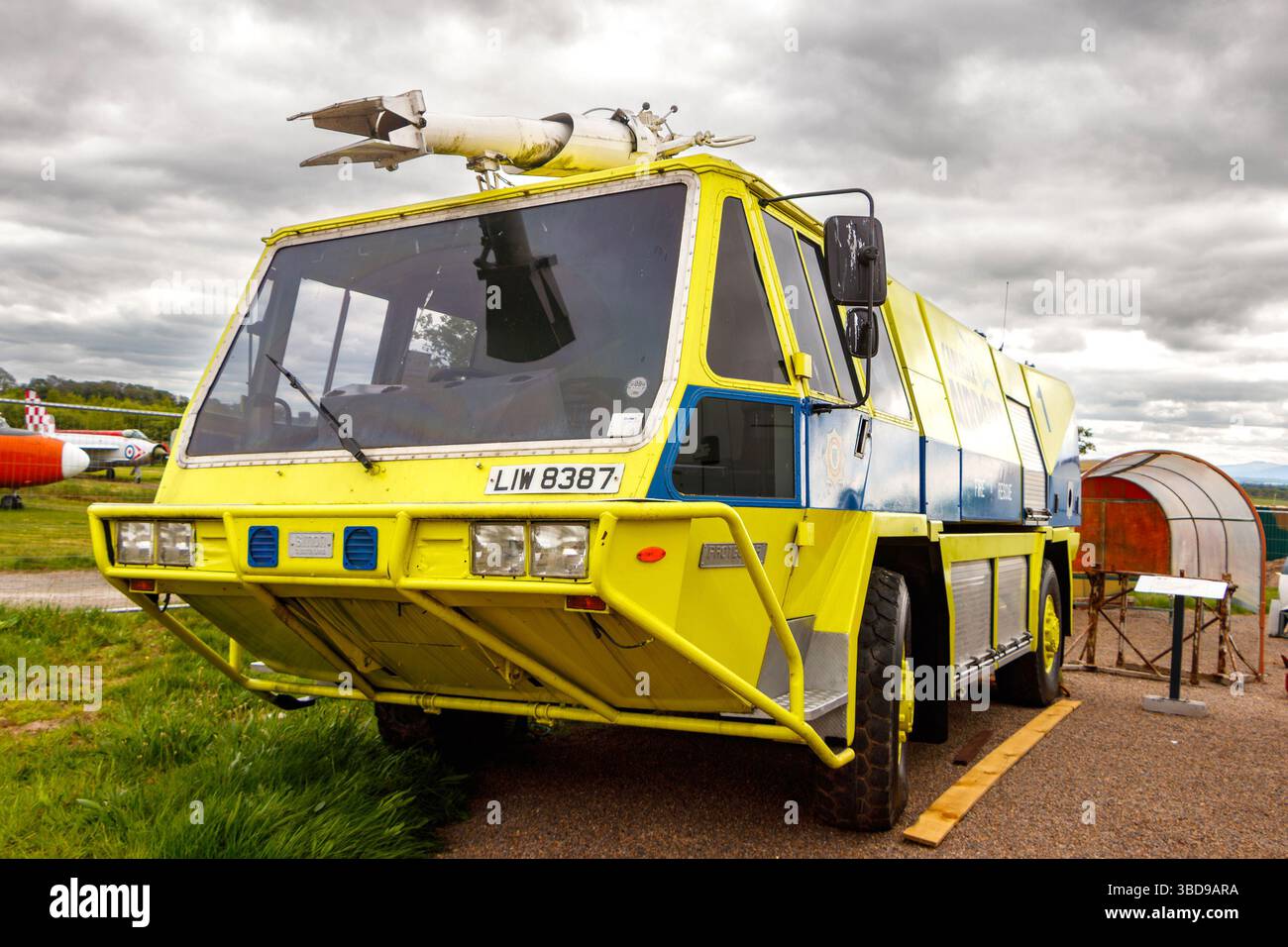 An einem bewölkten Tag parkt ein großer grün-gelber Flughafen-Feuerwehrwagen im Freien mit Start- und Landebahn und Flugzeug im Hintergrund Stockfoto
