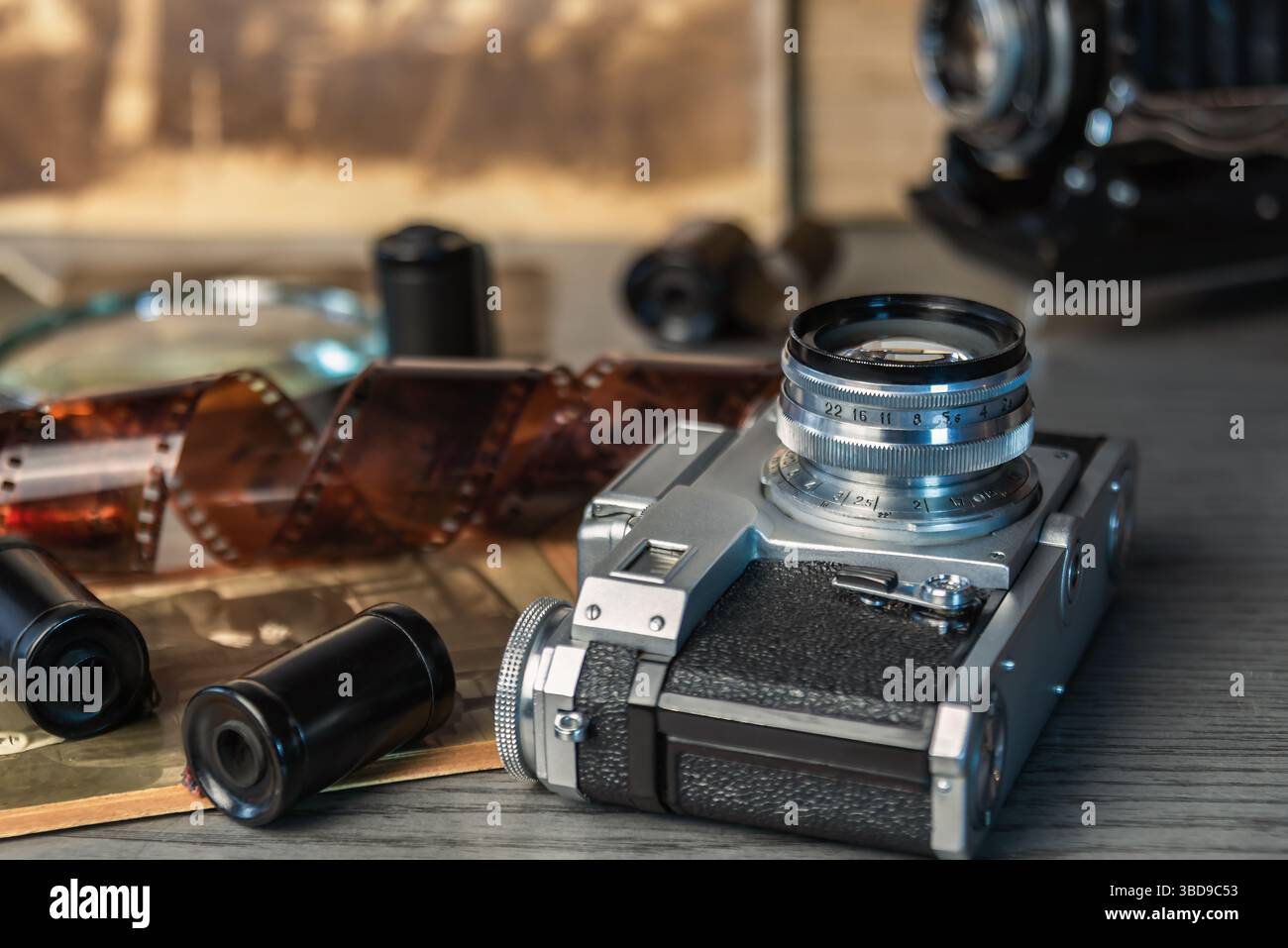 Die klassische Kamera liegt auf einem Holztisch, umgeben von alten Filmrollen und Fotografien, die eine nostalgische Atmosphäre der Fotografie hervorrufen. Stockfoto