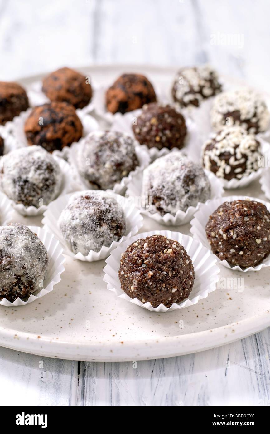 Verschiedene hausgemachte rohe vegane Energiebälle Cupcake Liner weiße Platte. Verschiedene Beschichtungen umfassen Kakaopulver Kokosnussflocken Nüsse. Gesunde vegane Süßigkeiten Stockfoto