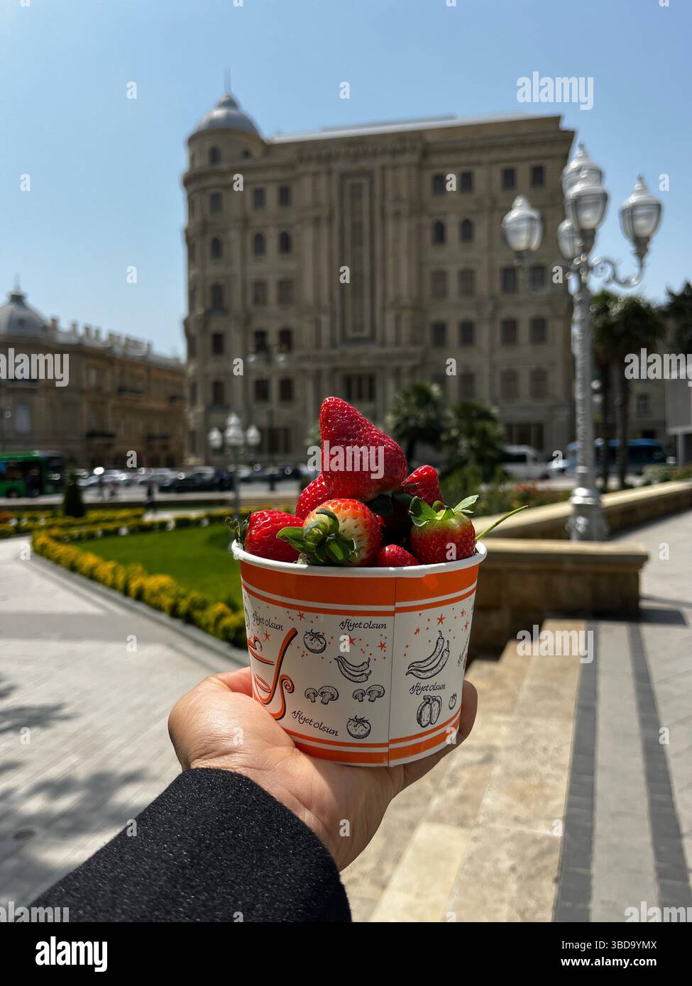 Eine Hand hält eine bunte Tasse mit frischen Erdbeeren auf einem Stadtplatz, mit einem großen historischen Gebäude und klassischen Straßenlaternen im Hintergrund. - Smartphone-aufgenommenes Stockfoto