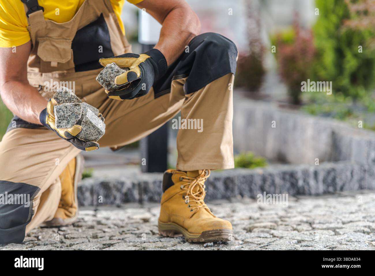 Ein Bauarbeiter kniet auf einem Steinweg und hält zwei Steine in der Hand. Er trägt Schutzhandschuhe und robuste Arbeitsstiefel, wobei er sich auf das konzentriert Stockfoto