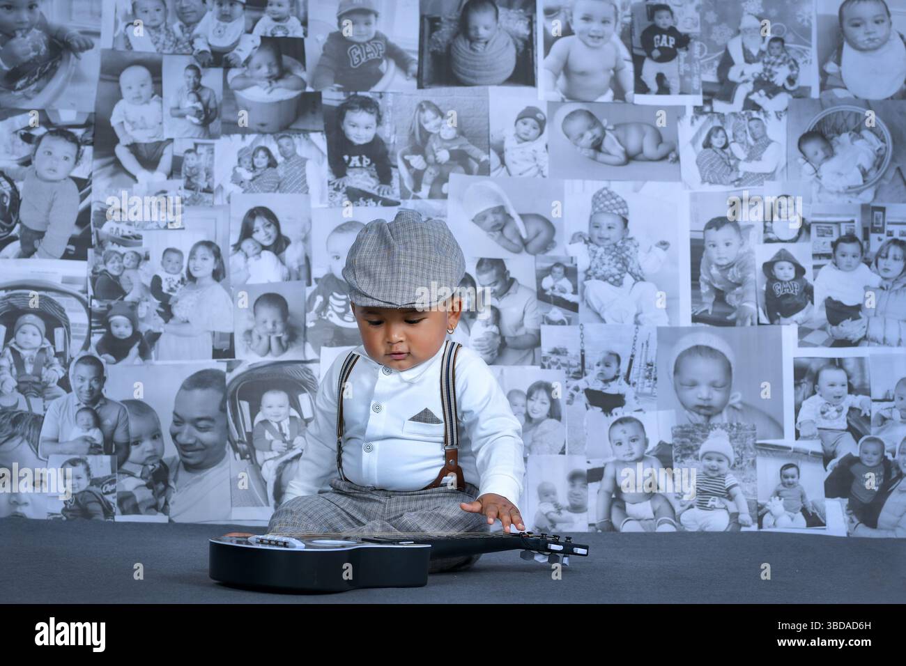 Der Junge im Vintage-Outfit spielt mit einer Spielzeuggitarre vor einer Collage seiner Kindheitsfotos während seiner ersten Geburtstagsfeier. Stockfoto