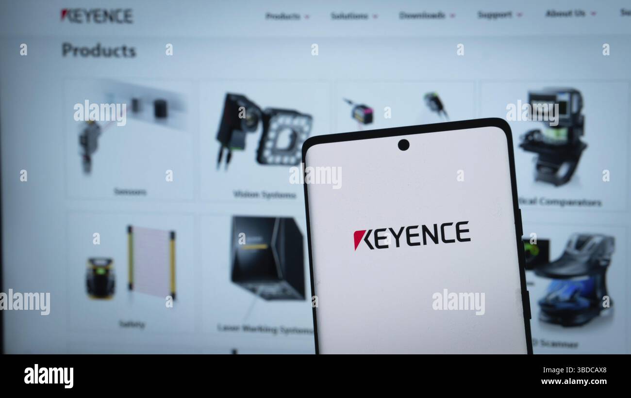 KEYENCE Logo auf einem Smartphone mit verschiedenen elektronischen Produkten dahinter Stockfoto