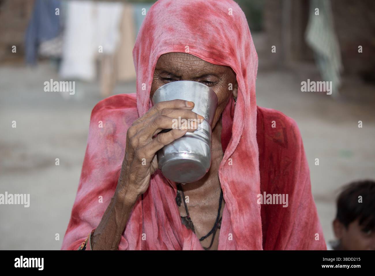 Ältere Frau in einem roten Schal, die Wasser aus einem Metallbecher in einem ländlichen Umfeld trinkt Stockfoto