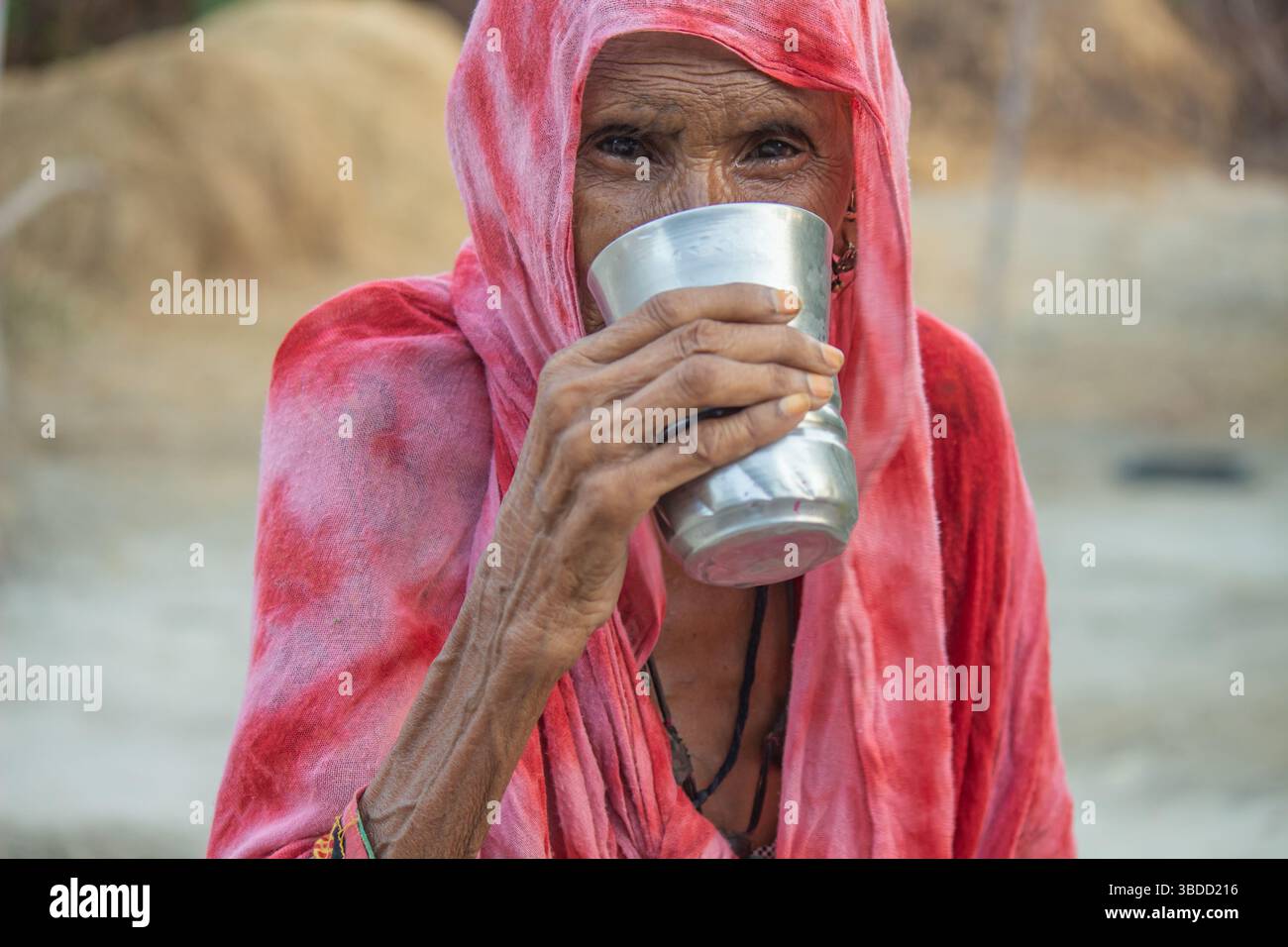 Ältere Frau in einem roten Schal, die Wasser aus einem Metallbecher in einem ländlichen Umfeld trinkt Stockfoto