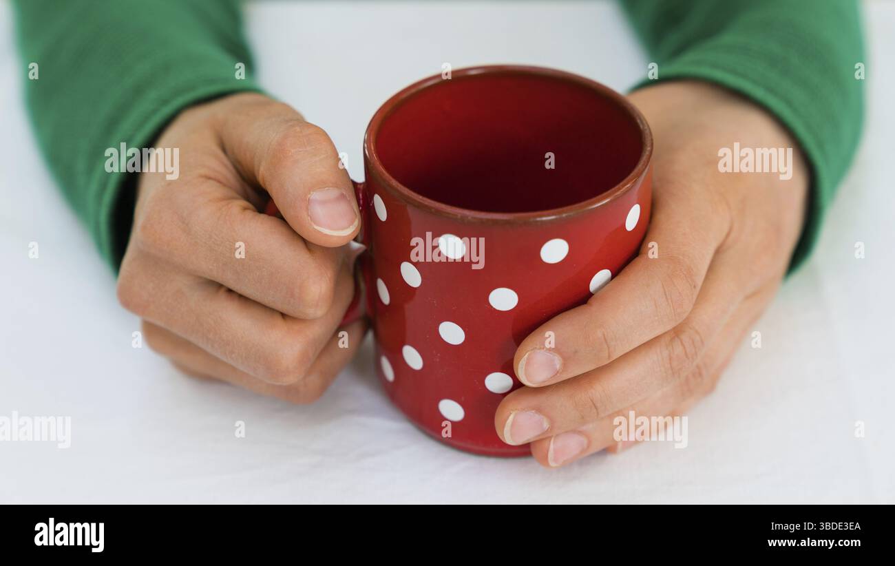 Zarte weibliche Hände einer Frau in einem hellen grünen Pullover holding Eine leere Kaffeetasse Rot mit weißen Tupfen auf einer weißen Tischdecke Stockfoto