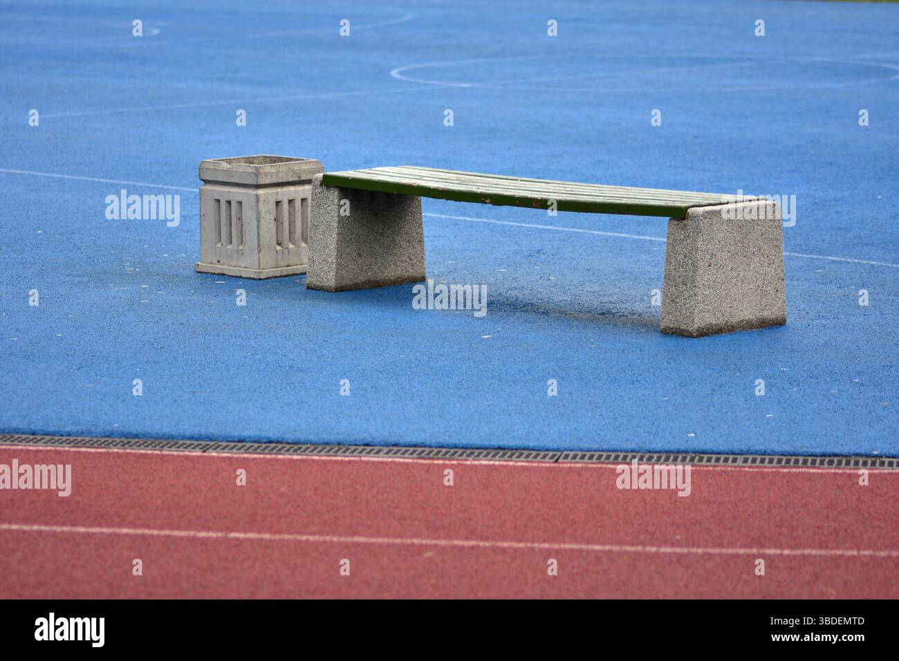 Blaues Sportfeld, rote Laufbahn und leere Bank mit Mülltonne bei bewölktem Tag. Stockfoto