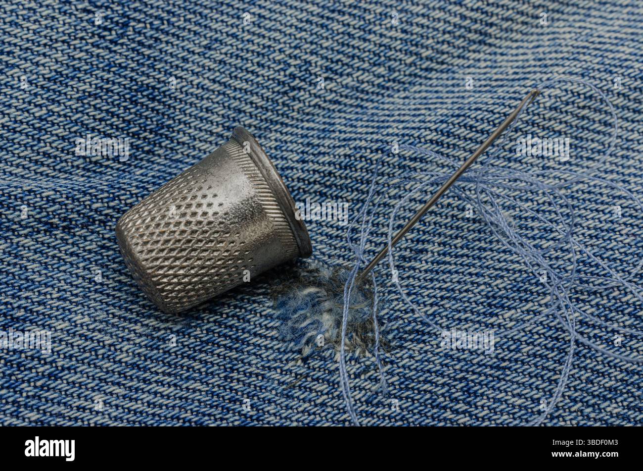 Nahaufnahme einer Metallkausche, einer Nähnadel mit Garn und einem Denim-Stoff mit sichtbaren Näharbeiten, die traditionelle Textil-Repai symbolisieren Stockfoto