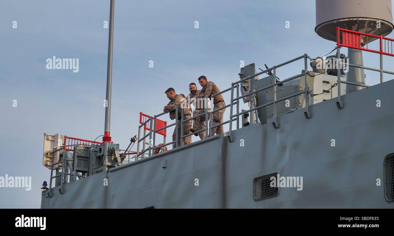 Spanien Malaga 1 28 2024 drei Soldaten, die aktiv auf einem Schiffsdeck unter einem teilweise bewölkten Himmel eingesetzt sind, umgeben von Marineausrüstung Stockfoto