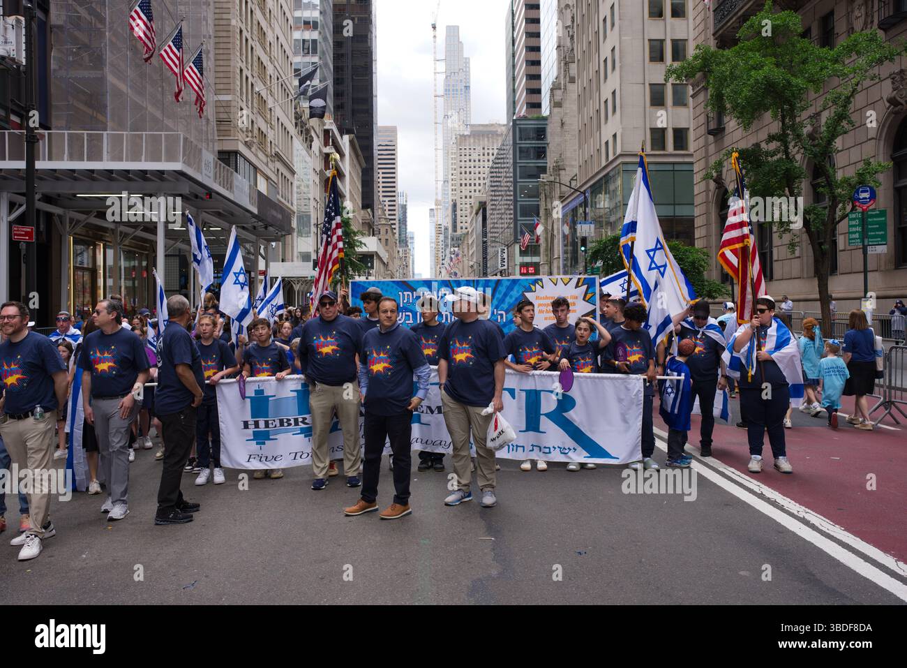 Eine Gruppe von Menschen, viele Kinder, marschieren in einer Parade eine Stadtstraße hinunter, die israelische und amerikanische Fahnen trägt. Sie halten ein Banner mit Hebräisch und Engländern Stockfoto
