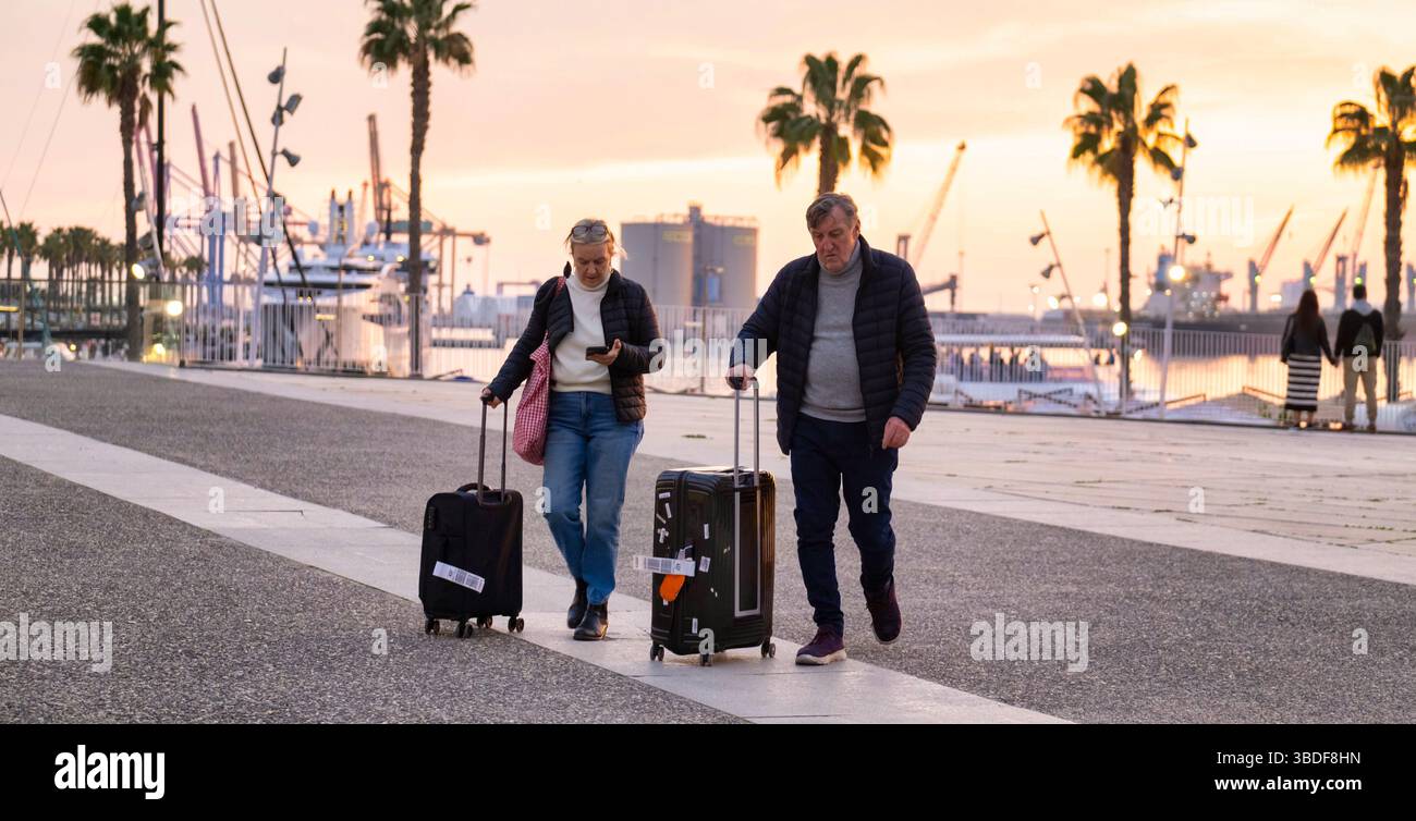 Spanien Malaga 1 28 2024 reisendes altes Paar mit Koffern in der Nähe der ruhigen Uferpromenade bei Sonnenuntergang, geschmückt mit Palmen, Booten und Hafenkranichen Stockfoto